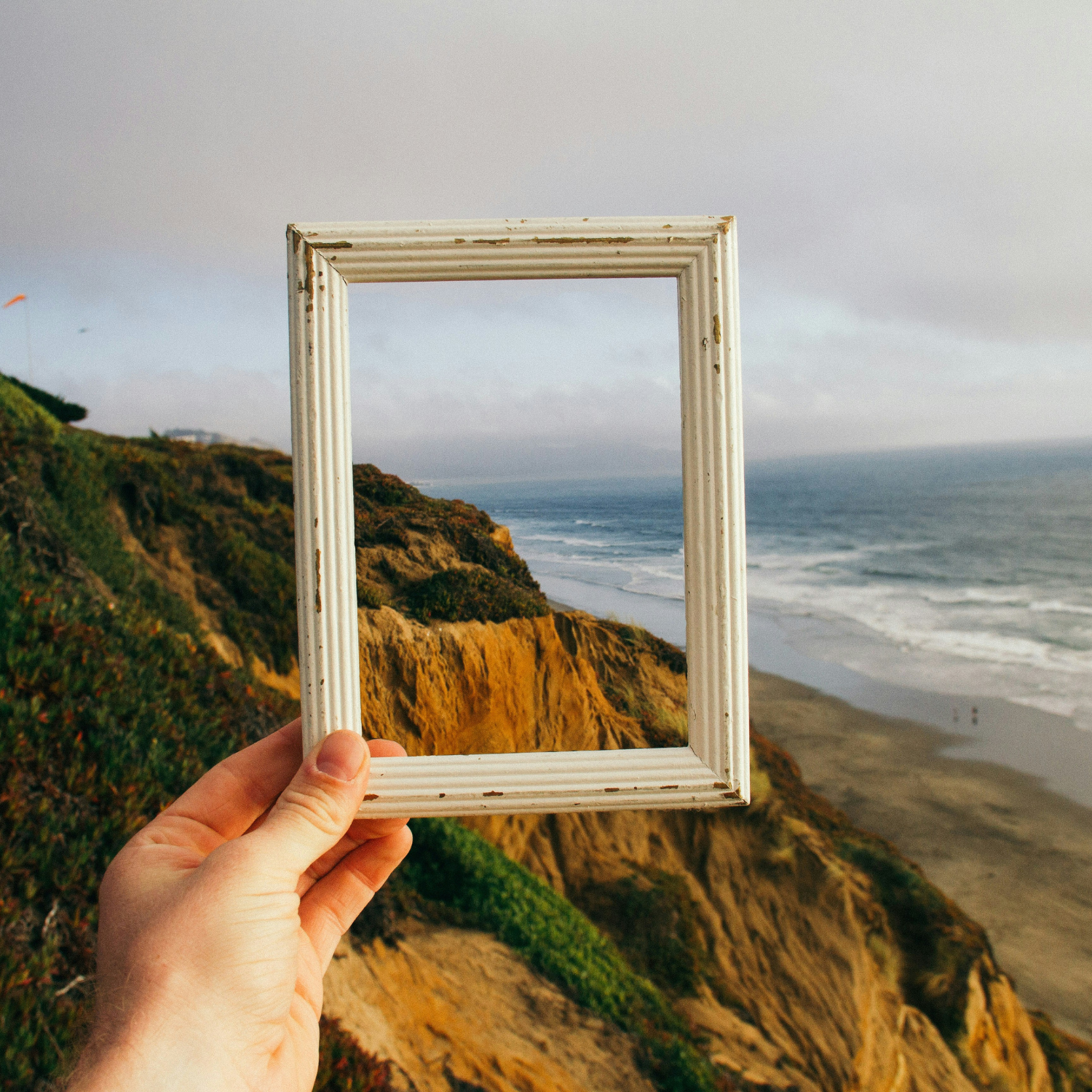 An image of a hand holding a square wooden picture frame in front of a picturesque landscape as an allegory of decision framing in eCommerce. Through the frame, you can see a view of the ocean, sandy cliffs, and lush greenery.